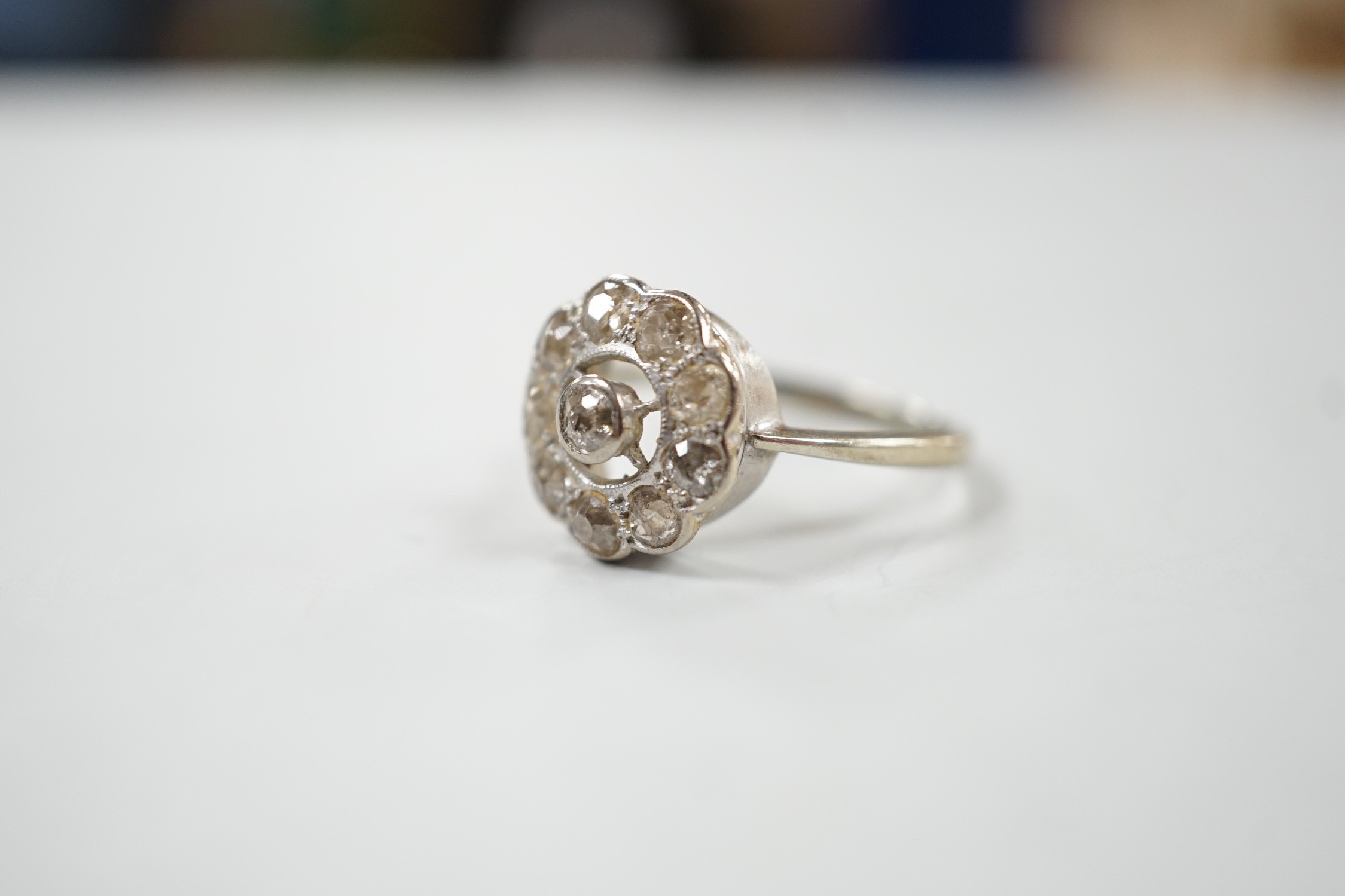 A 1920's white metal and ten stone millegrain set diamond flower head cluster ring, size P/Q, gross weight 3 grams.
